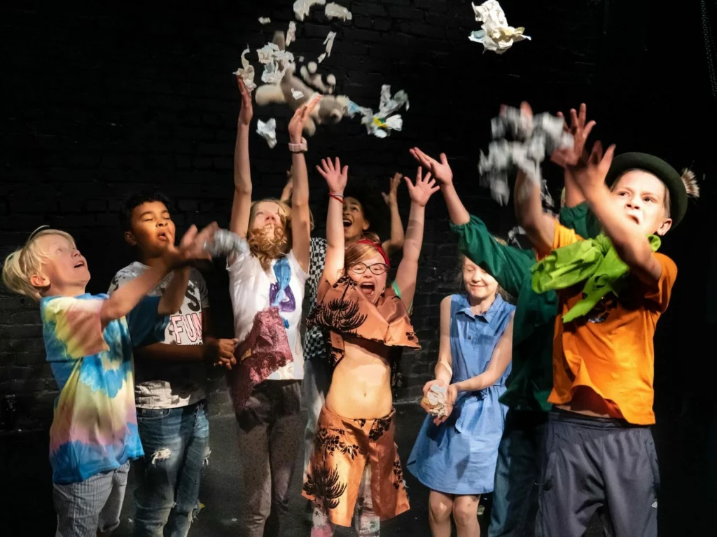 Flera glada barn från teaterskolan Konfetti står på scenen och kastar konfetti upp i luften.Useat iloiset lapset teatterikoulu Konfetista seisoo lavalla ja heittävät konfettia ilmaan.