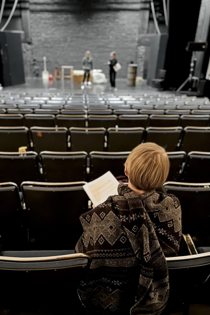 En regissör sitter i Karelias sal och ser på två personer som står på scenen.Ohjaaja istuu Karelian salissa ja katsoo kahta näyttämöllä olevaa henkilöä.