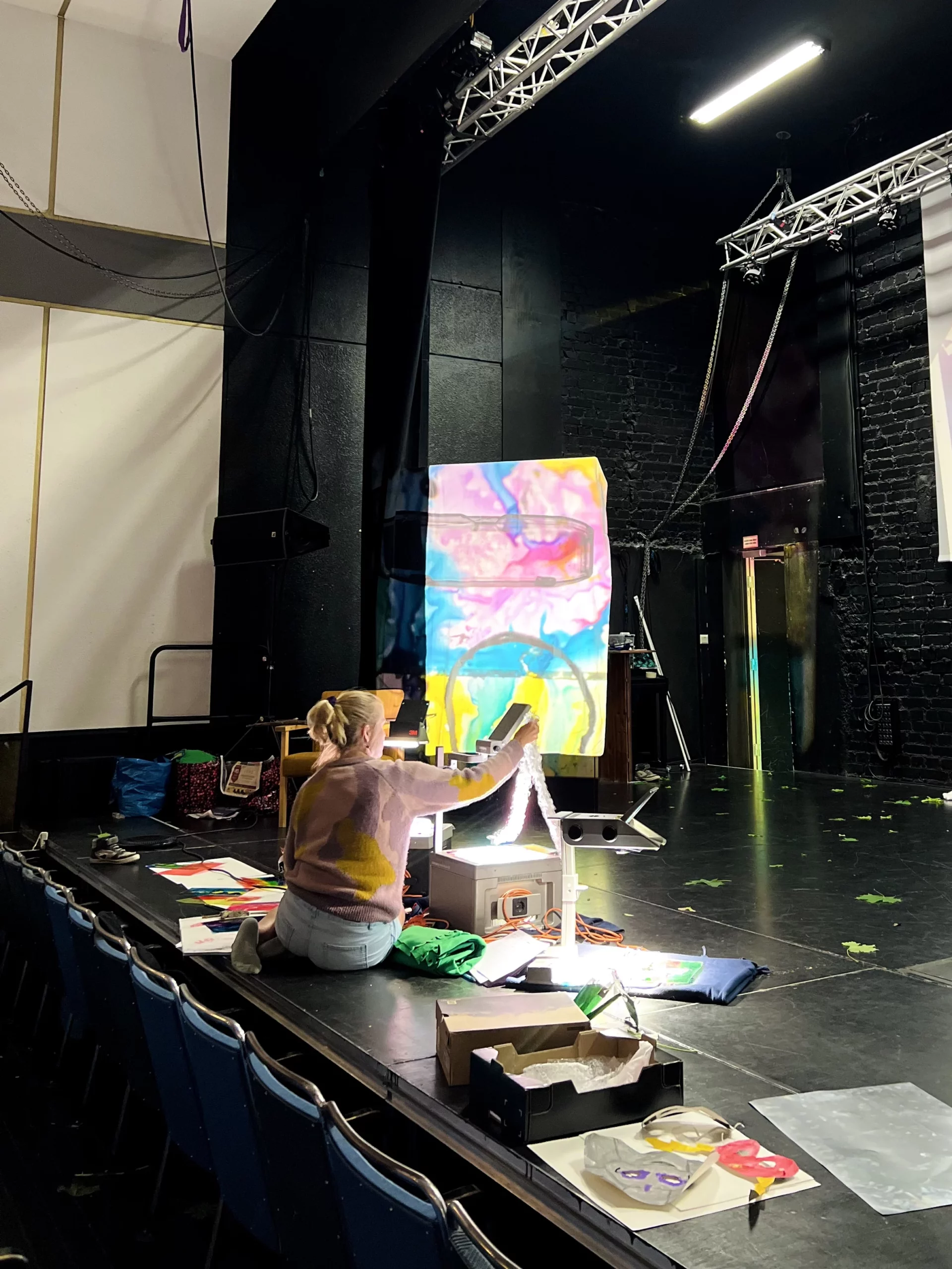 En konstnär sitter på Karelias scen och visar upp ett färgglatt konstverk med hjälp av en OH-apparat.Taitelija istuu Karelian näyttämöllä ja esittää värikästä taidetta piirtoheittimen avulla.